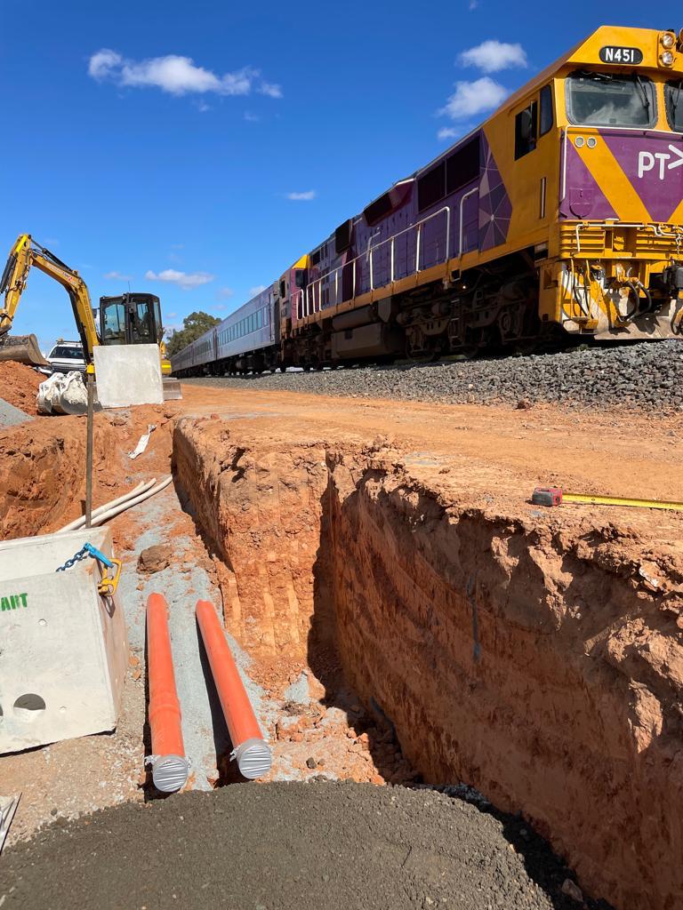 A purple and yellow train travels on the tracks alongside a construction site. A deep trench is dug beside the tracks, with orange pipes visible. A small excavator is parked near the trench under a clear blue sky.