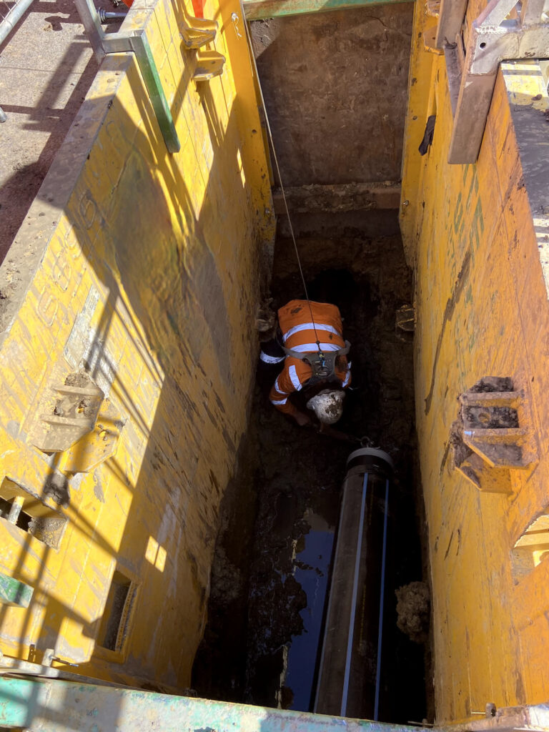 In the heart of Victoria's infrastructure, a worker in an orange safety vest and helmet is crouched in a deep trench, surrounded by yellow protective barriers, performing maintenance on a large underground pipe as sunlight casts shadows across the site.