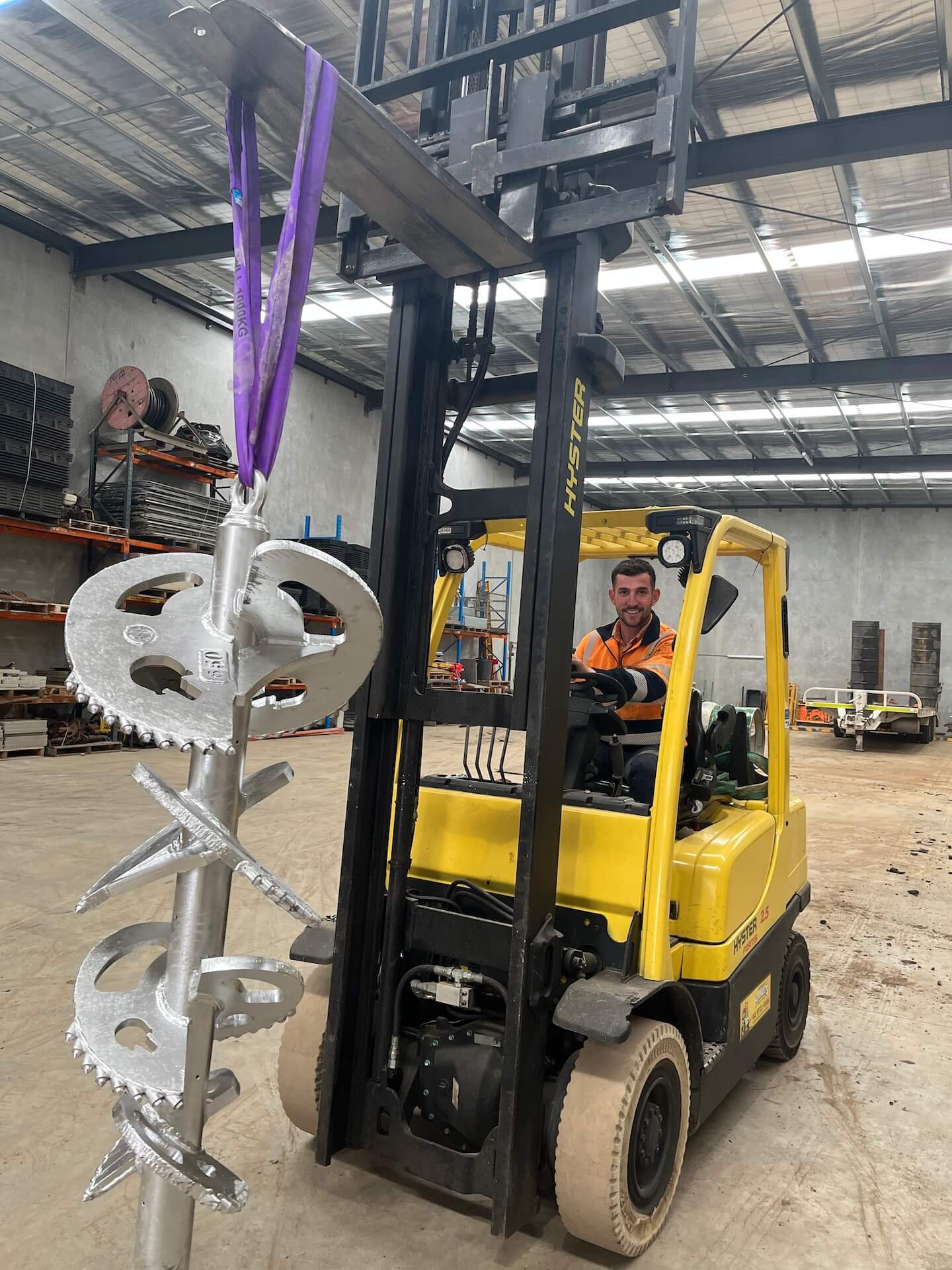 A man in a high-visibility shirt skillfully operates a yellow forklift within the warehouse, lifting a large silver metallic auger with purple straps. Shelves and construction materials, forming part of the site's comprehensive infrastructure solutions, are visible in the background.