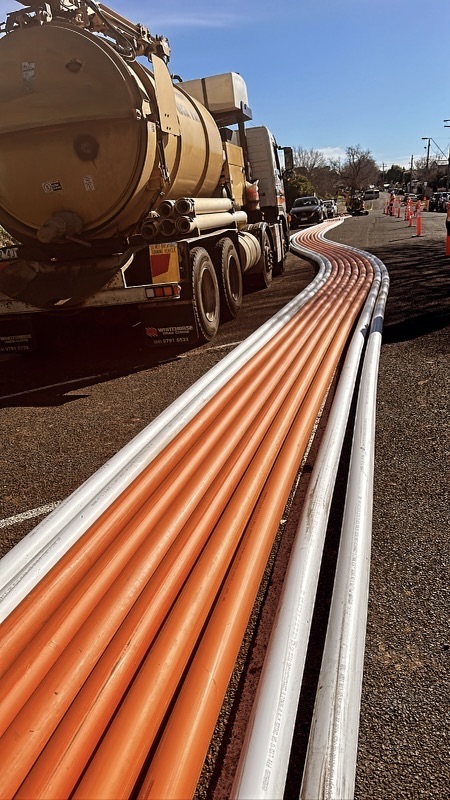 A truck equipped with a vacuum excavation system is parked on the road, providing infrastructure solutions. Long orange and white cables extend down the street, lined with traffic cones under a clear blue sky.